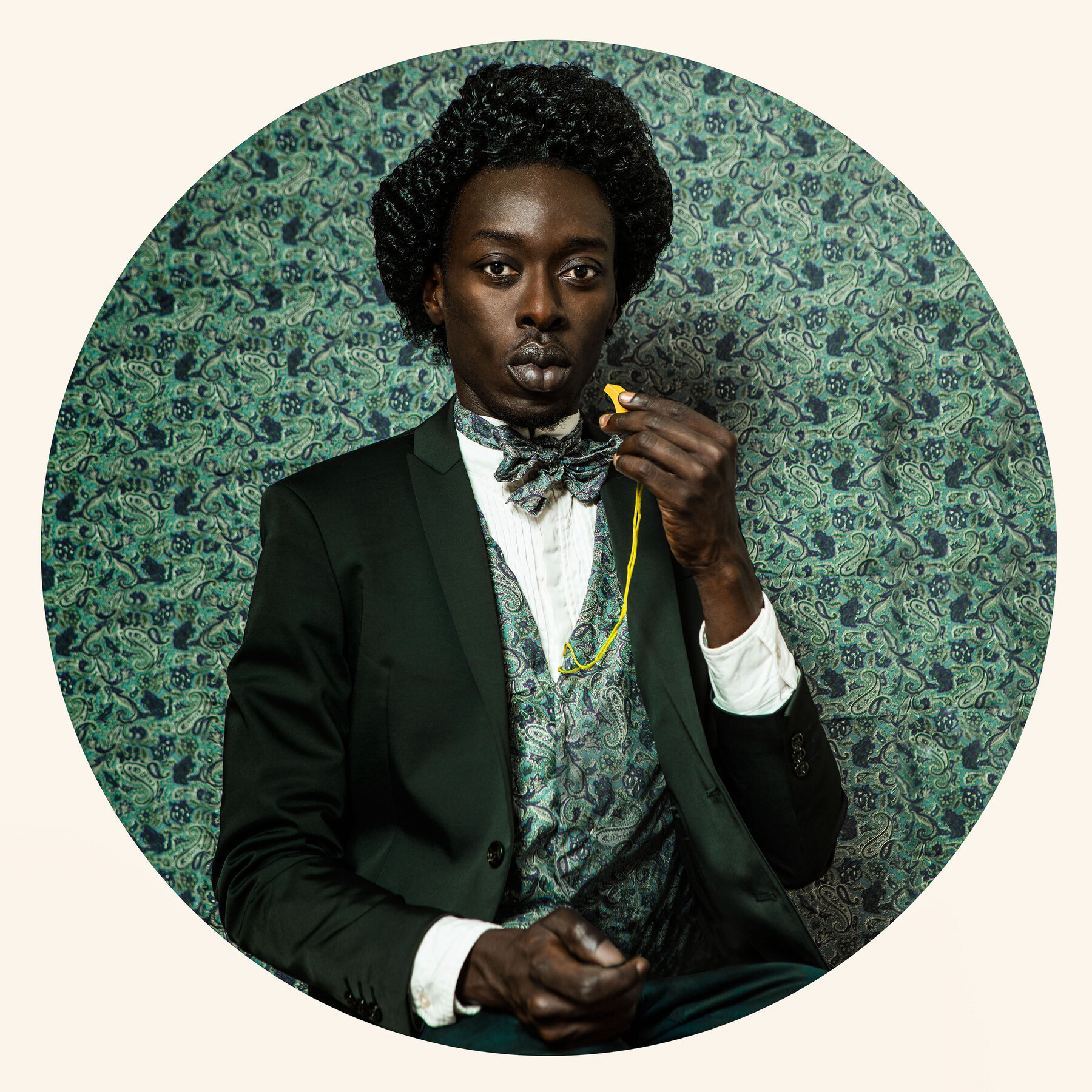 Omar Victor Diop wearing a three-piece dark green suit with a white shirt and holding a yellow whistle close to his lips. His green paisley vest and bow tie match the background behind him.