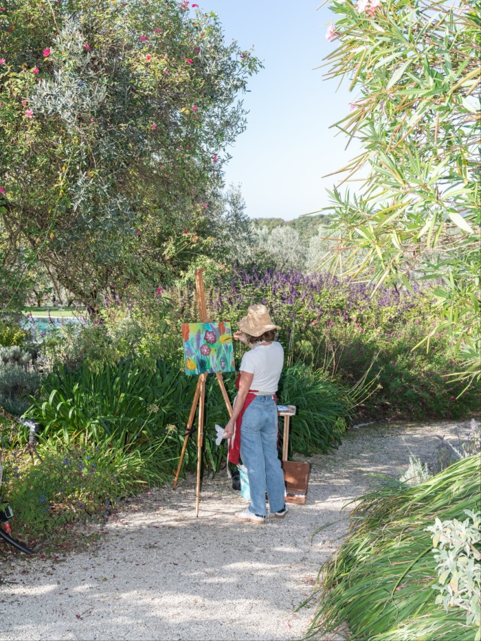 A woman stands on a garden pathway, wearing a straw sun hat and painting at an easel