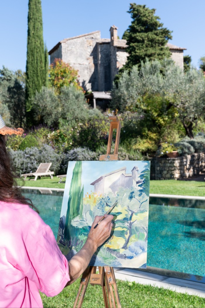 A view of a woman from behind, at an easel, painting a historic building with a swimming pool in the foreground
