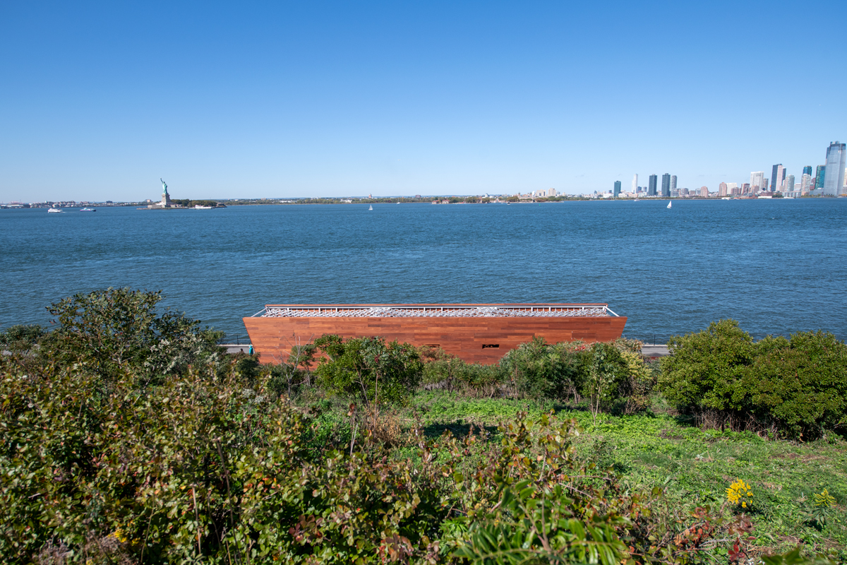 Aerial view of a wooden sculpture that resembles a ship with the New York harbor, including the Statue of Liberty, in the background. 