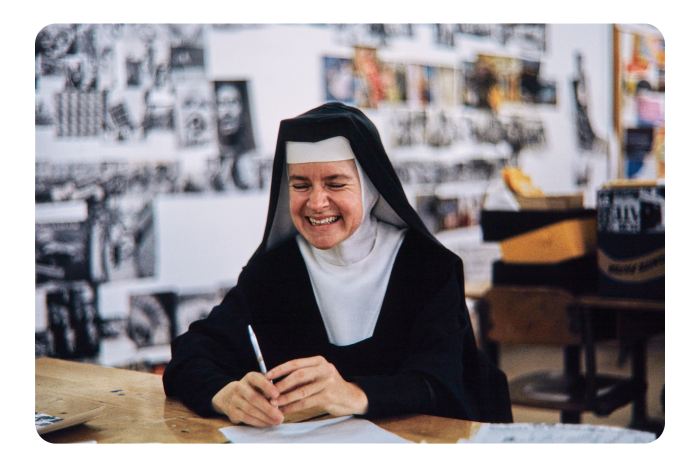 A picture of Sister Mary Corita in her nun’s headpiece and habit. She is smiling at the camera and holding a pen