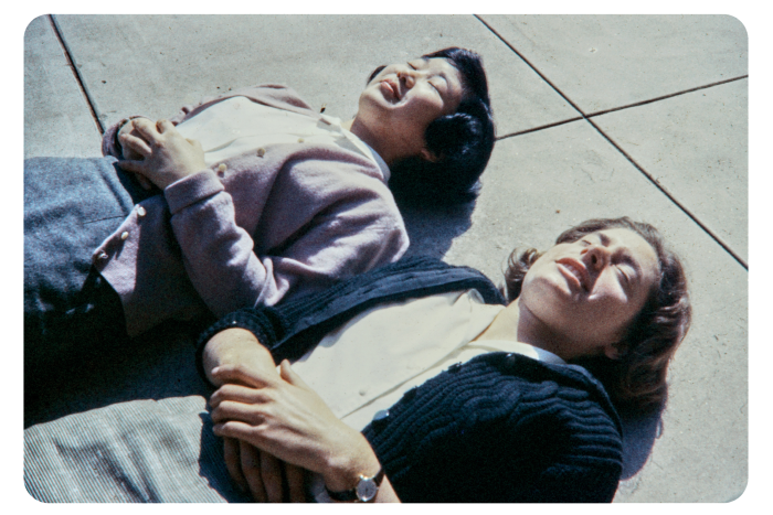 Two women lie smiling, side by side, sunbathing. Strangely, they are fully clad and appear to be lying on concrete slabs