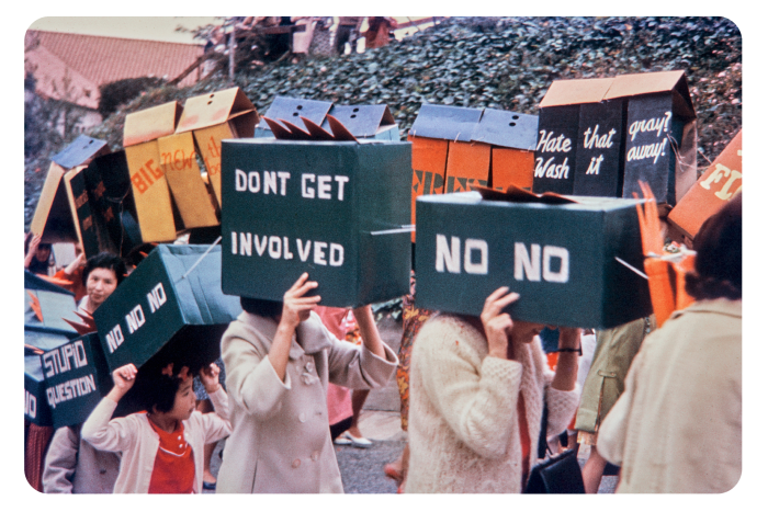 A photograph of women on a protest, possibly against US involvement in Vietnam. Instead of placards they are carrying cardboard boxes painted with slogans, including, ‘No, no’, ‘Don’t get involved’, ‘Stupid question’