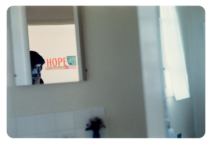 A picture of a mirror in a bathroom. Reflected on the back wall we glimpse the top of Corita’s head in her nun’s habit, her hand holding a camera, and a sign with the word ‘Hope’ in big orange letters 