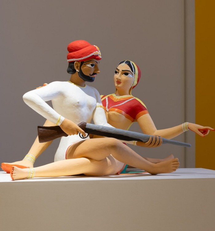 In a sculpture, a man in a turban holding a rifle is intertwined with a woman on a plinth