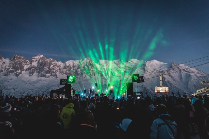 Green light beams up from a stage at night. Crowds of people watch, and in the background there are snowy mountains