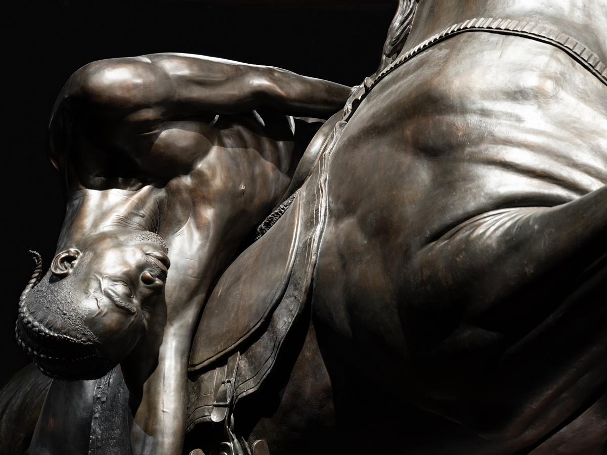 In a realistic bronze statue, a Black man with braids lies slumped over a horse's saddle.