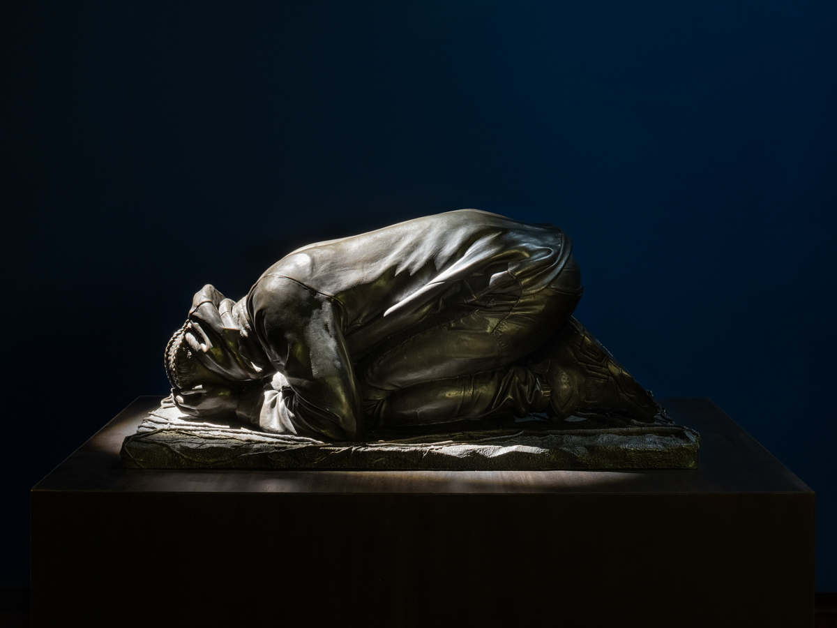 A bronze sculture of a Back man with braids, wearing a hoodie and sneakers, crouched over with his head in his hands.