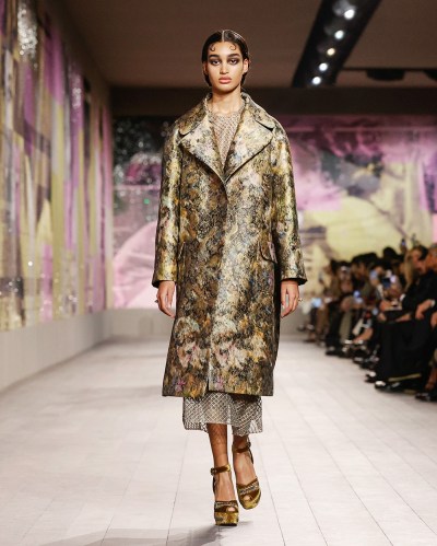 A person with light brown skin has curled baby bangs and is walking down the runway in a metallic coat. Behind them is a pink and yellow collage with archival imagery.