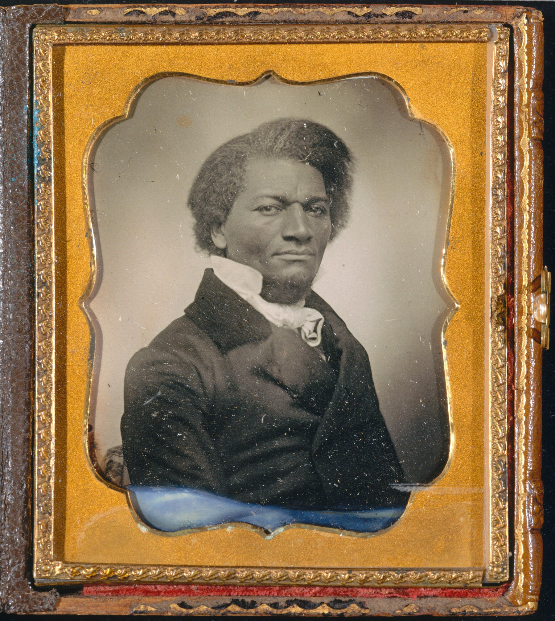Frederick Douglass posing for a seated portrait, in mid-19th century attire. The portrait is black-and-white and encased in a gold frame.