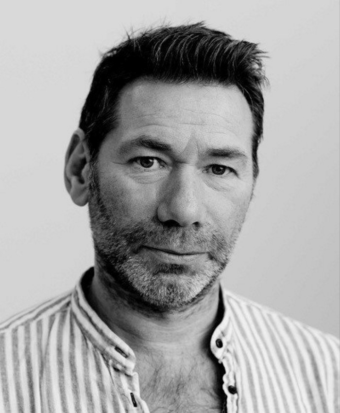 A black-and-white photo portrait of Mat Collishaw in a striped open-necked shirt