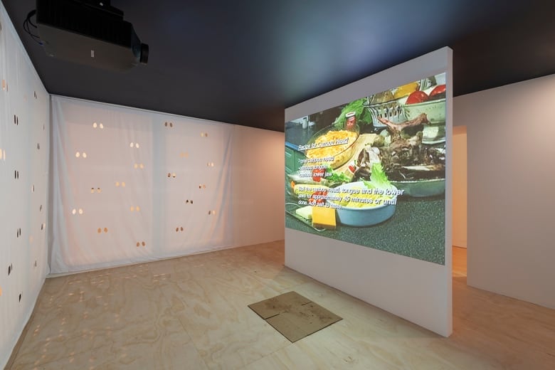 Photo of an art installation in a white-walled gallery. Visible: Walls covered with cut-outs, reminiscent of the eyes in a sheet-ghost Halloween costume. A video is screened on one partition in the middle of the room. The imagery is of counter covered with food. Text for a recipe appears overlaid on the image.