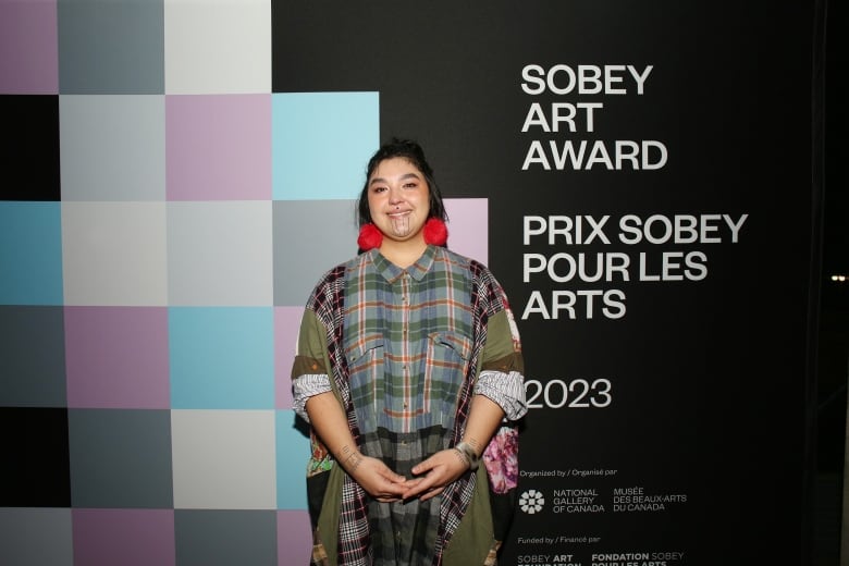 Medium shot of the artist Kablusiak. They stand smiling in front of a black step-and-repeat wall branded by the 2023 Sobey Art Award.