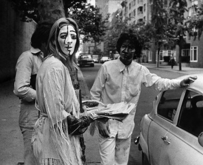 A woman with a painted face holding papers beside two men and a car.