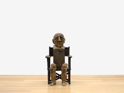 A squat figure seated in a chair without arms. His hands rest on the chairs sides and on his legs.
