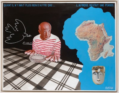 A man in a striped red Breton shirt seated a table where rolls are arranged so that they form his hands. He sits beside a drawing of a dove, an image of the African continent, and a mask.