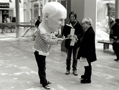 A person wearing a giant mask resembling the head of an old man signing a sheet of paper in an atrium.