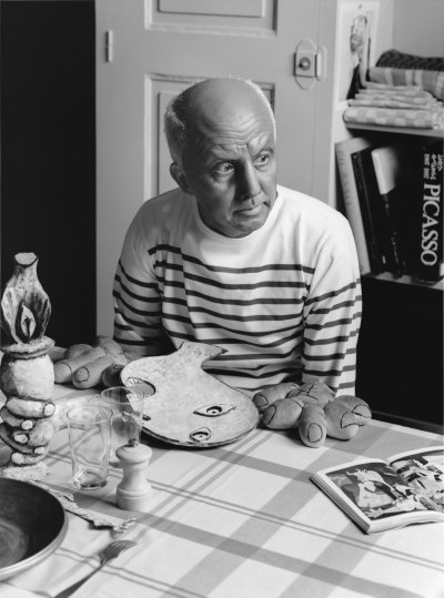 A black-and-white photograph of a man in a striped shirt seated a table. A plate resembling a crying woman's head is set before him, along with rolls made to look like his enlarged hands. A book lies open to a page showing a print of a painting.