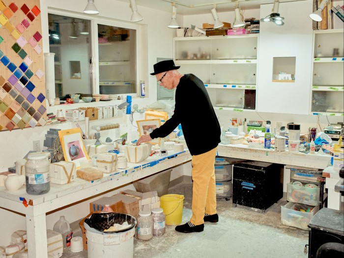 A man in a black cardigan pottering around his bright studio