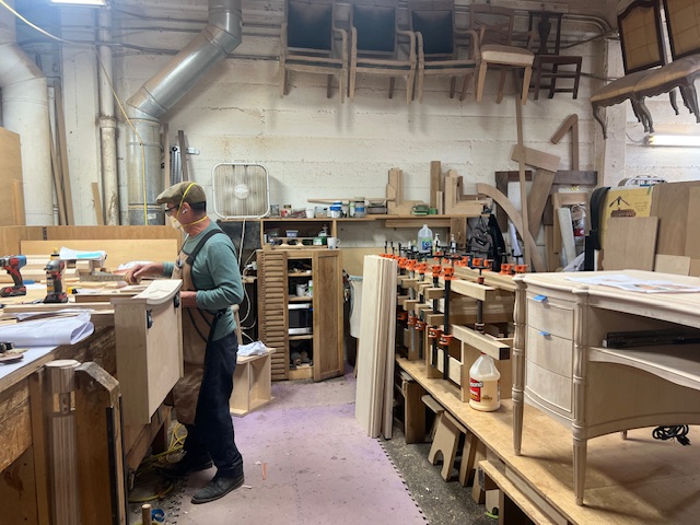 A man with a mask and a hat on works on sanding wood in a workshop. 