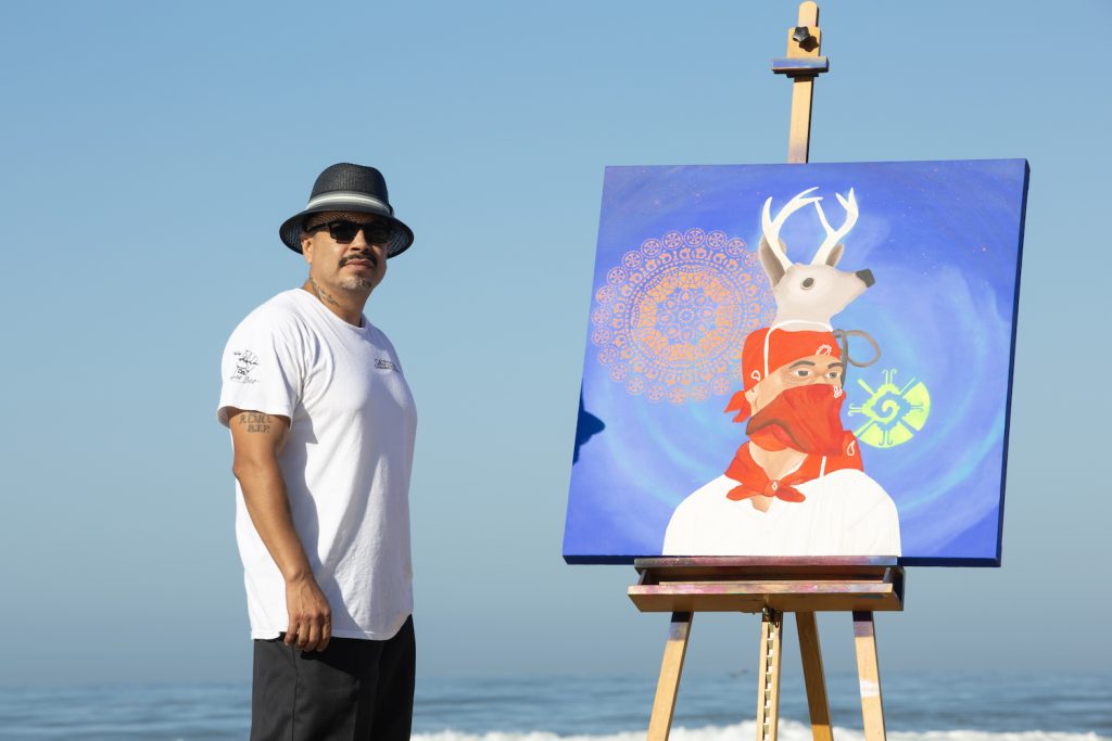 Tijuana artist Javier Salazar Rojas standing next to his painting of a man wearing a bandana and a reindeer hat
