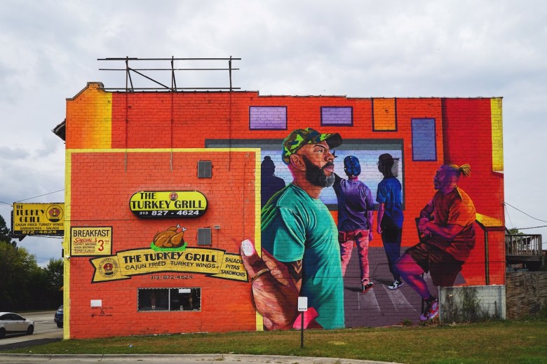 Red brick wall with a mural of a Black man wearing green looking to the right. Four other Black men are lined up at a cashier’s counter. In the foreground, a hand is holding a “menu” — it’s the building’s hand-painted signage, which reads, “The Turkey Grill. 313-827-4624. Breakfast special. Turkey sausage, hash browns, toast & jelly. Cajun fried turkey wings. Salads, pitas, sandwiches.”