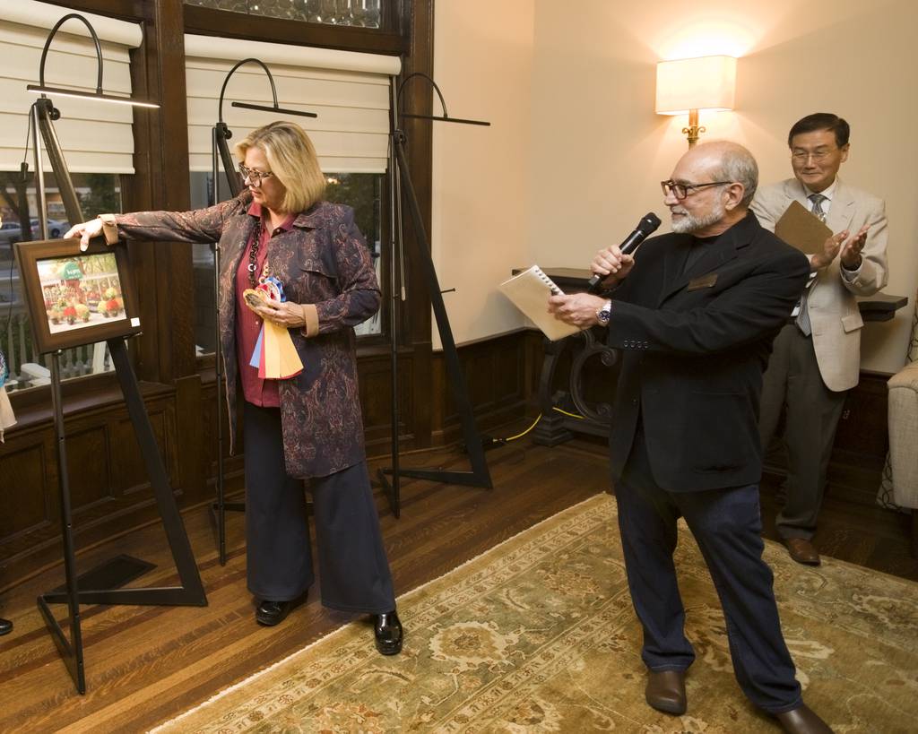 Thomas Balsamo, of the Barrington Cultural Commission, right, speaks as Janette Tepas, from the Barrington Area Artists Association, left, places a ribbon on the artwork that placed third in the Plein Air art event in Barrington. The winning pieces were on display at the event reception held  Oct. 14, 2023 at Barrington's White House in Barrington