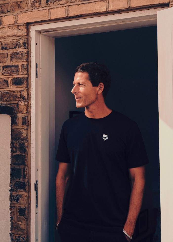 A man with short dark hair, wearing a black T-shirt and trousers, stands in a doorway, hands in pockets