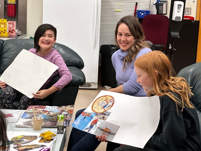 Two smiling children sit holding pieces of large paper they've made collages on.  A woman watches and laughs with them.