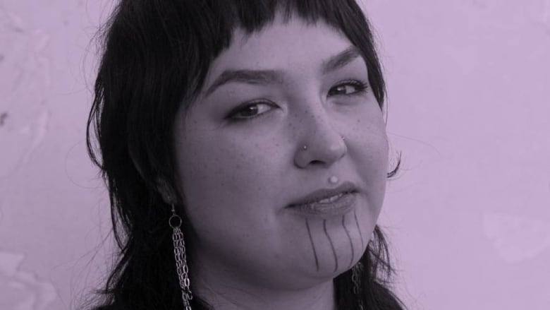 Headshot of the artist Kablusiak. A black-and-white photo that's been tinted lavender. The artist, who uses they/them pronouns, has dark straight hair that's been cut into a mullet. They smile at the camera. Their chin is tattooed with four vertical lines and they wear earrings and facial piercings.