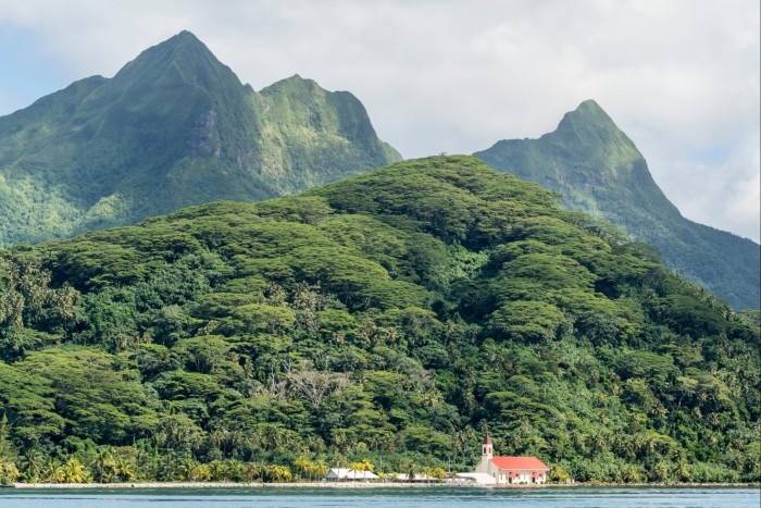 A view from the sea of a building at the foot of a forested hillside, with mountains rising up behind it