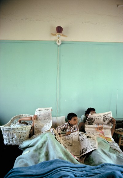 A Black child and two unseen people with newspapers in front of them on a bed.