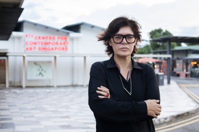 A woman with thick-rimmed glasses with her arms crossed before a building.