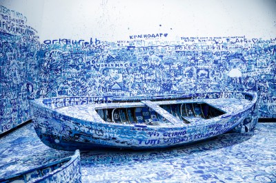 A rowboat tipped on its side in a gallery. Both the walls, the floor, and the boat itself are covered in scrawled white and blue text.
