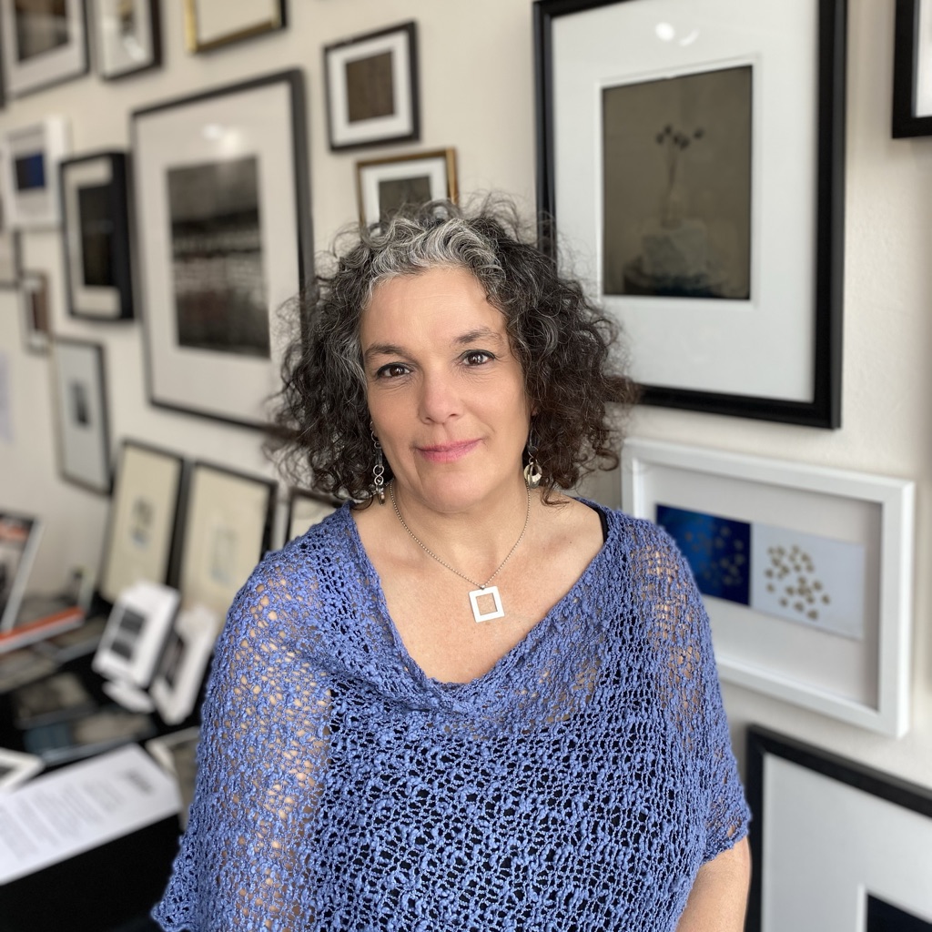A woman in a blue knit shawl and dark curly hair, gray in front, stands in a stuio in front of famed photos.
