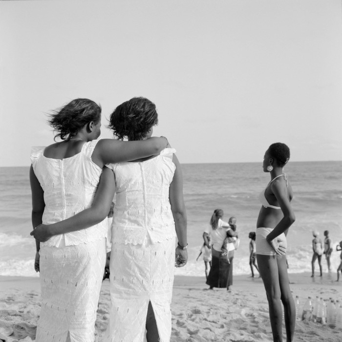 Two women wearing white dresses hug each other while standing on a beach