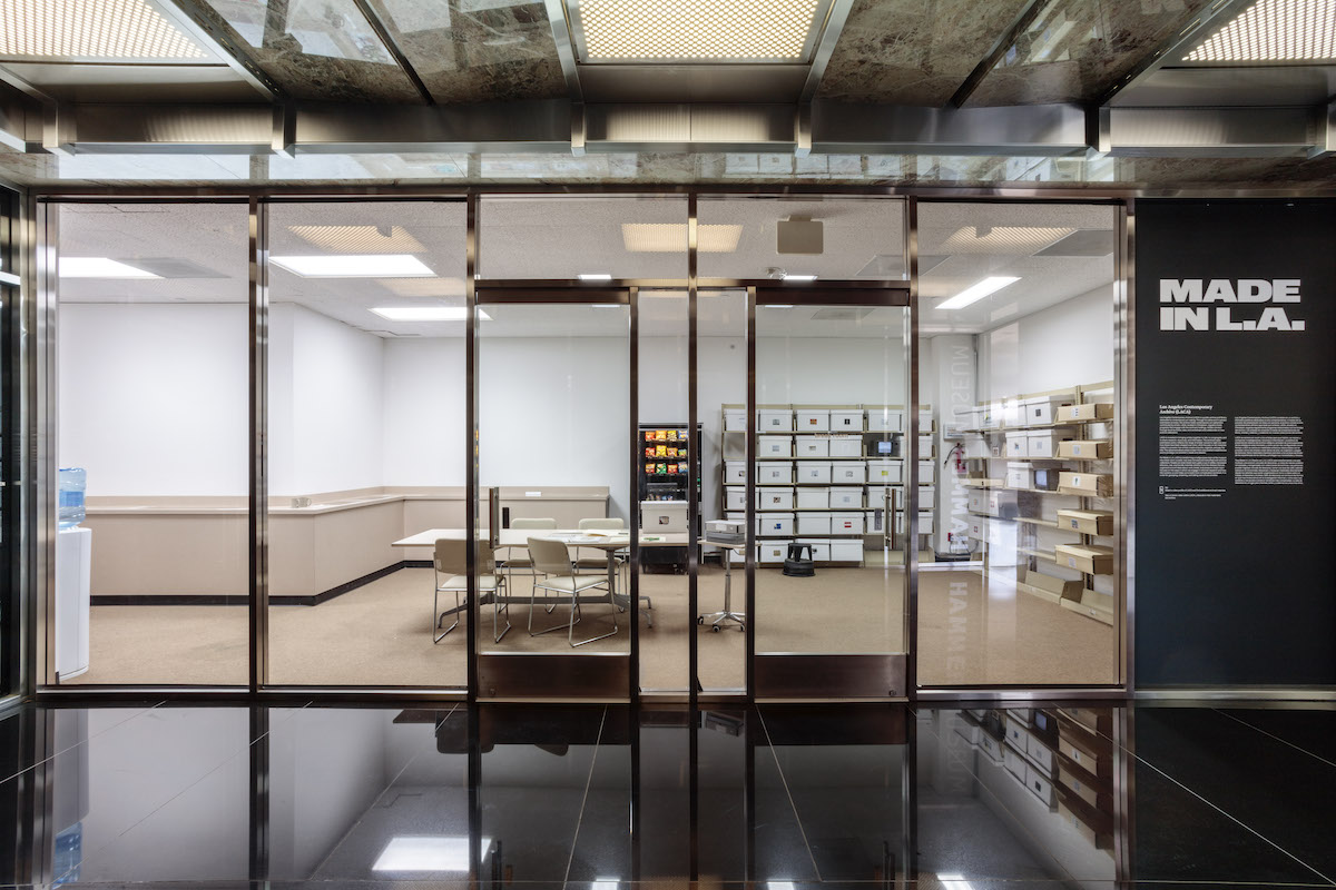 A glassed-in room with an office table, some chairs, and many boxes of files in a shelving unit. A vending machine and a water cooler can also be seen.