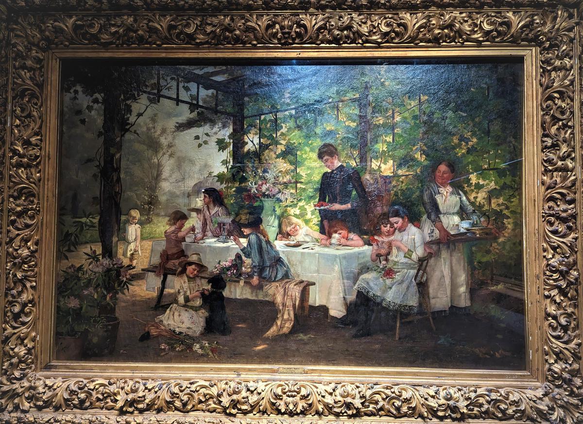 ‘Children’s party’, a 20th century painting by German artist Heinrich Schwiering shows many playful children and adults gathered around a table under a pergola.