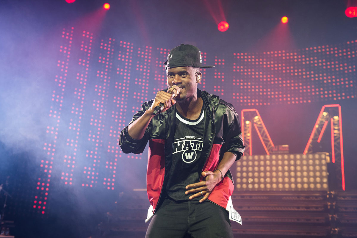 A Black man wearing a baseball cap and leather jacket holds a microphone as he sings on stage (hip-hop)