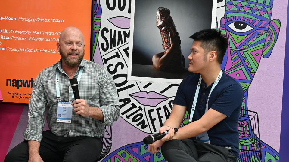 Two men sit in front of a picture of a sculpture shaped like a penis filled with antiretroviral pills