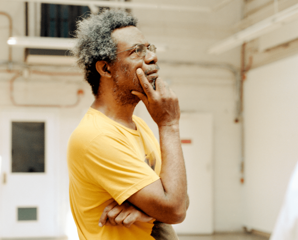 A Black man in a yellow T-shirt with one hand to his chin.