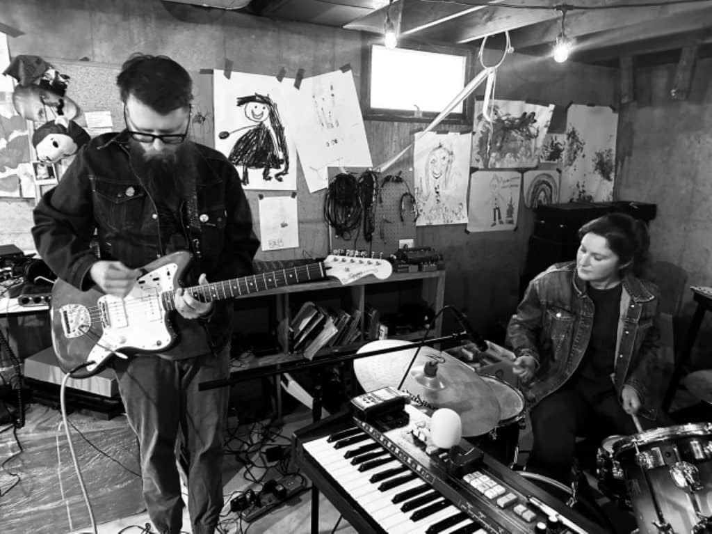 A black-and-white photo  of rock duo Red Pants playing in a practice space. Jason Lambeth plays guitar while standing and looking down to the left of the image. Off to his right is drummer Elsa Nekola. A keyboard is visible at the bottom of the image, slanted towards Lambeth's waist. Both Red Pants members are in dark clothing. A series of Lambeth's visual art adorns the wall behind them. 