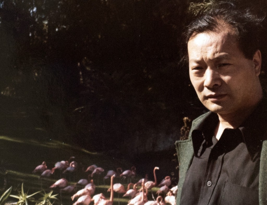 A color photograph of the Chinese American artist Walasse Ting in front of flamingoes at the wildlife park Parrot Jungle in Miami, February 1977. Photograph by Natalie Ting. From the Estate of Walasse Ting.