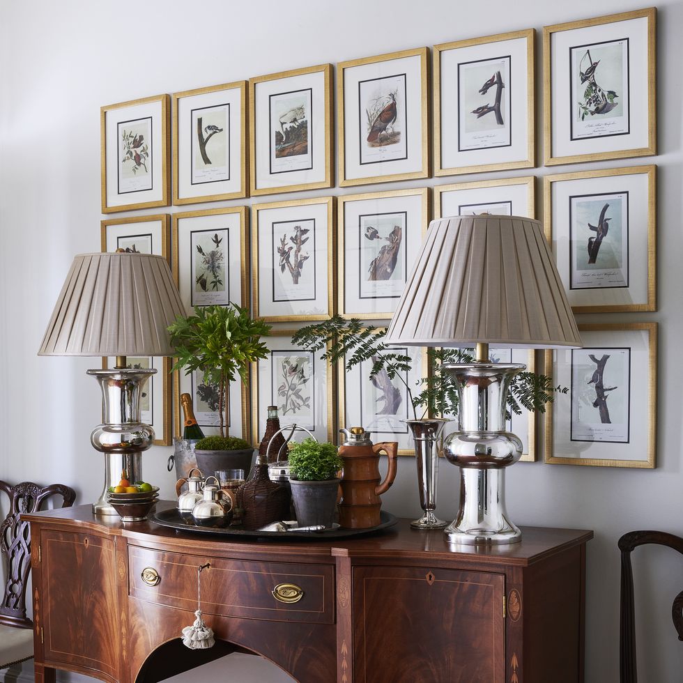 a series of antique nature prints hangs above a buffet with a pair of mercury glass lamps