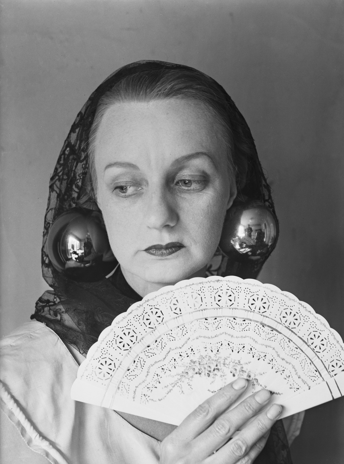 A woman with a head covering who wears giant reflective globe-like earrings. She holds a fan before her.