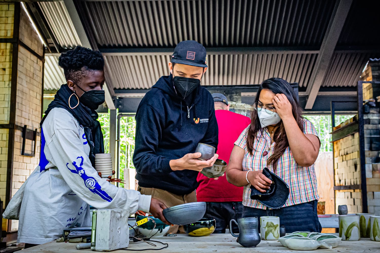 Three people wear face masks and stand over a table where they are looking at various ceramic pieces.