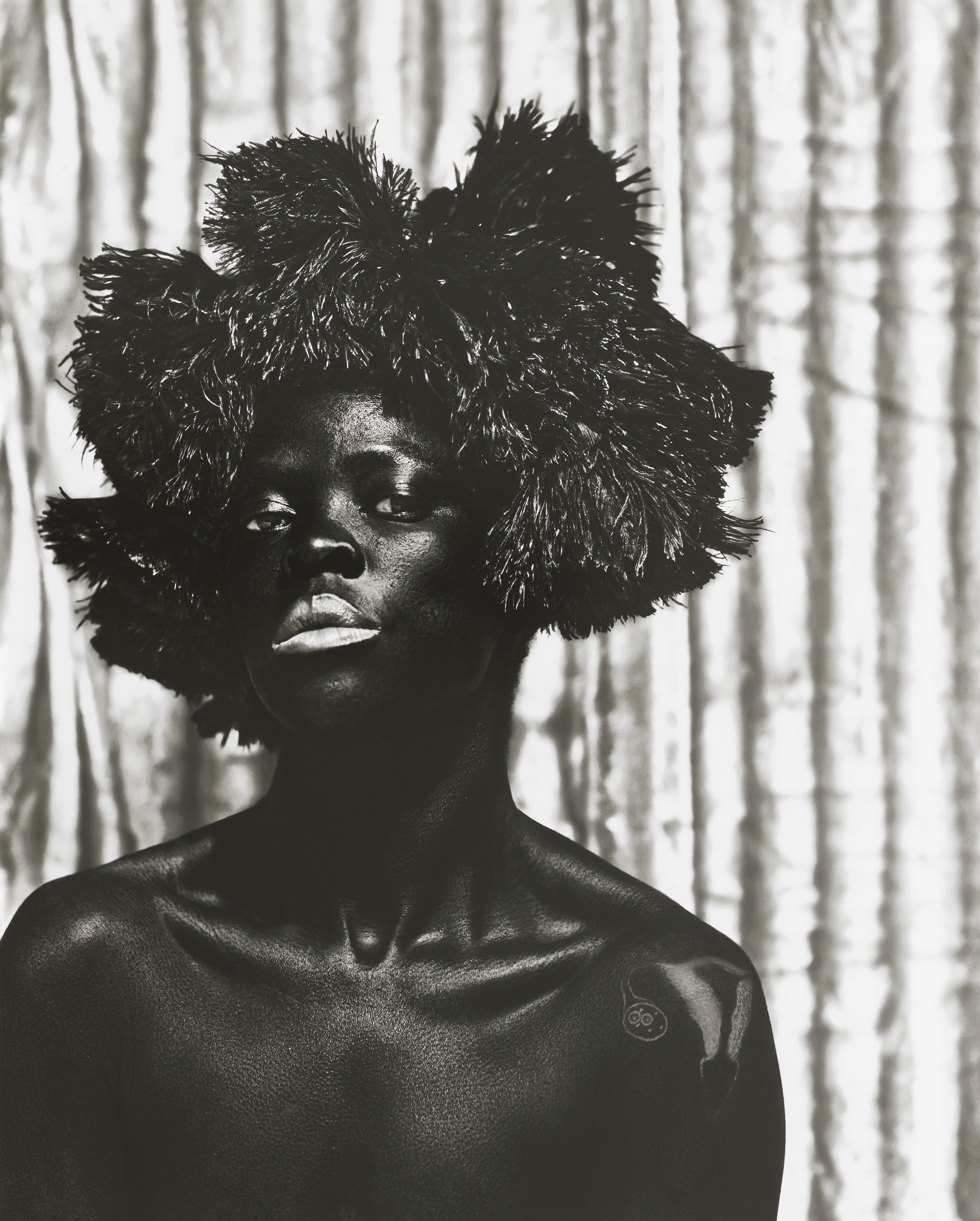 Black and white image of figure posing with spiky headdress against vertical stripe backdrop