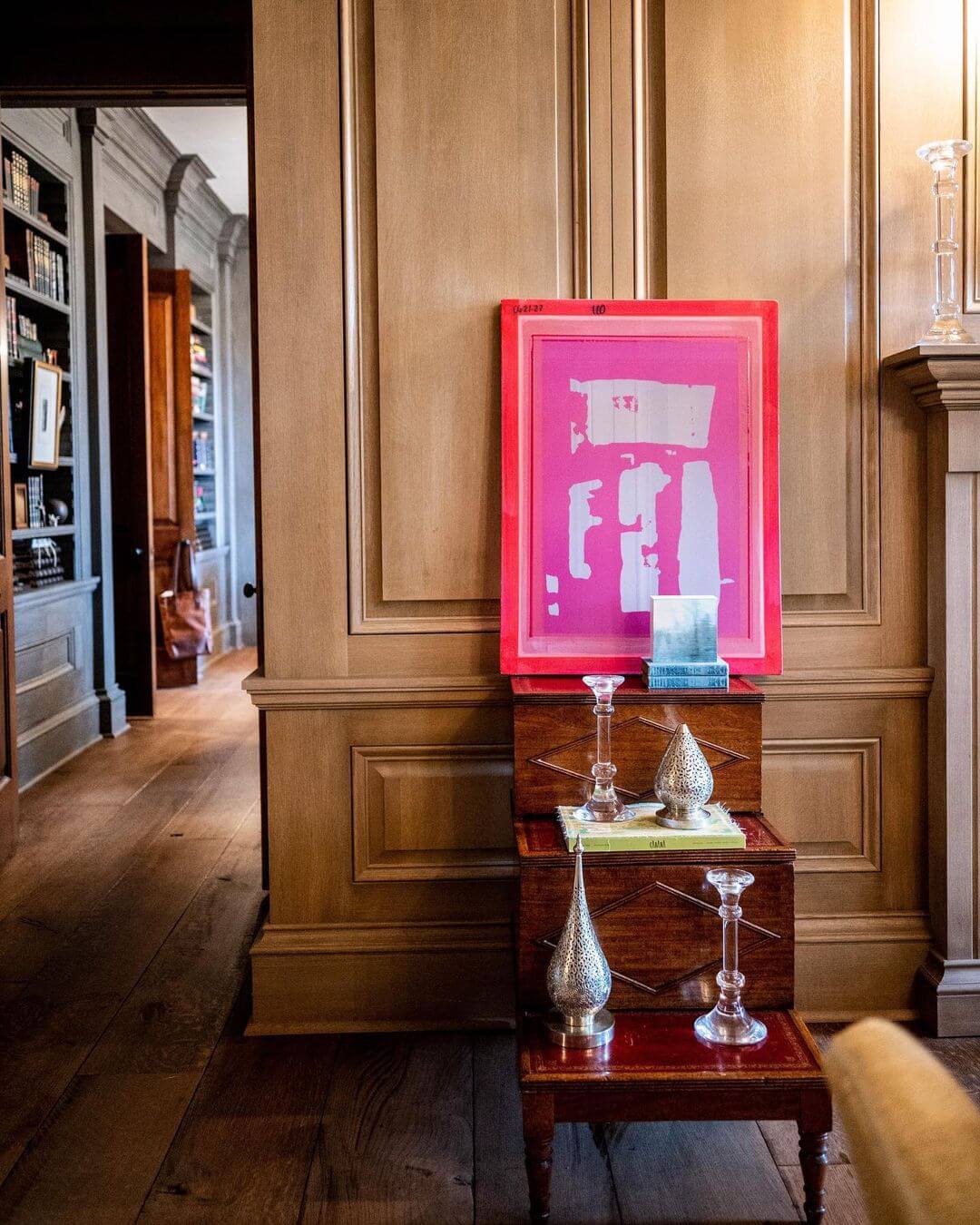 An elegant room with wooden paneling featuring a neon pink silkscreen art piece by Laura Deems on a small wooden table with decorative glassware.