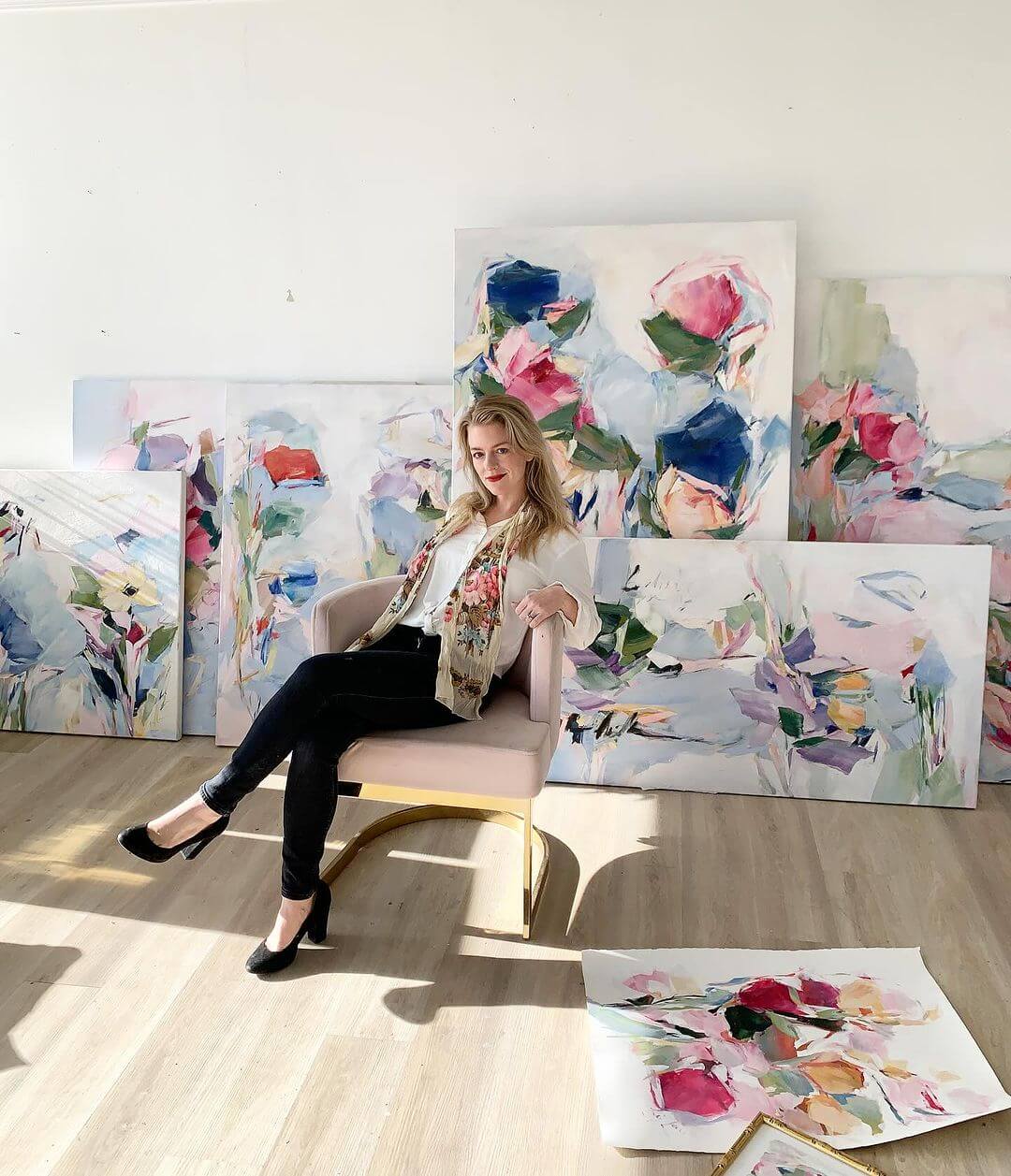 Liz Lane sitting on a chair in a bright studio surrounded by colorful floral paintings.