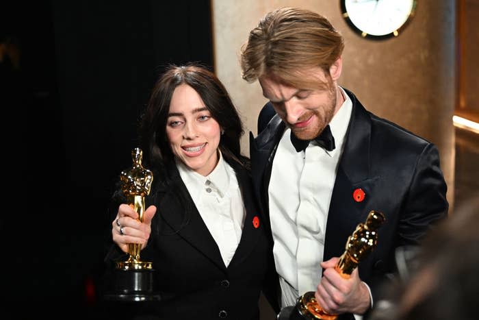 Two people smiling holding Oscar trophies, one in a white shirt and bowtie, the other in a tuxedo